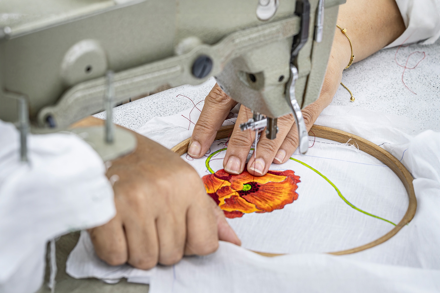 Mains brodant un coquelicot rouge sur un lin blanc tendu dans un cerceau, sous la tête d'une machine